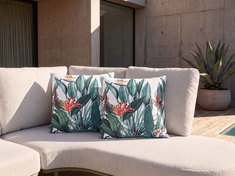 Zwei quadratische Zierkissen mit tropischem Pflanzenmuster auf einem hellen Outdoor Sofa vor einer modernen Betonfassade.