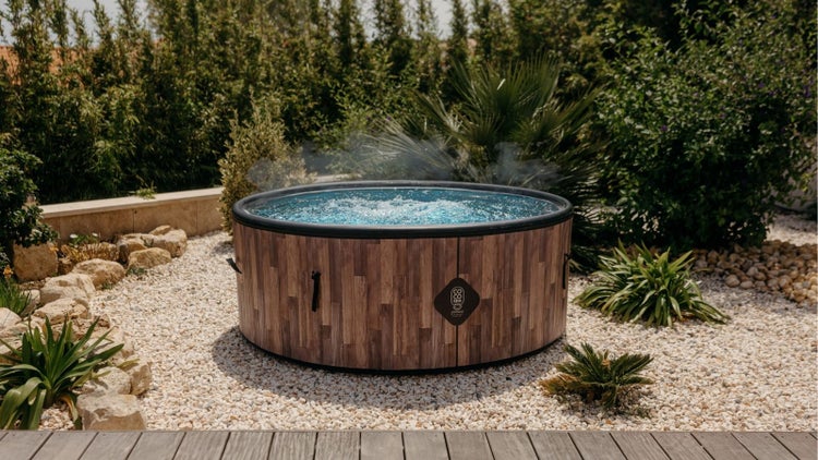 Runder, aufblasbarer Whirlpool mit Holzoptik-Verkleidung und dampfendem Wasser in einem Garten mit hellem Kies und grünen Pflanzen.