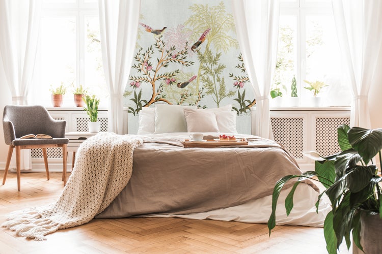 Helles Schlafzimmer mit botanischer Fototapete mit Vögeln und Bäumen, Bett mit beiger Bettwäsche, grauem Sessel und Fischgrätparkett.