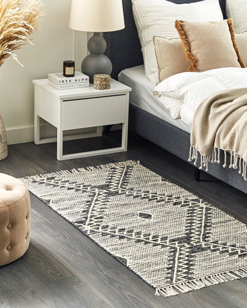 Gewebter Teppich mit geometrischem Muster und Fransen in einem Schlafzimmer mit Bett, Nachttisch und Hocker auf Holzboden.