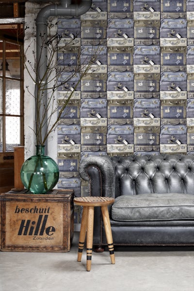 Szene mit einer Wandtapete mit Koffermotiv, einem grauen Ledersofa und einem Holzhocker