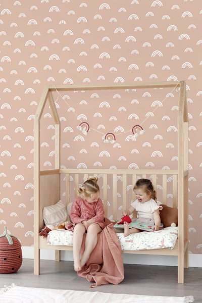 Zwei Kinder sitzen in einem Holzbett vor einer Wand mit Regenbogenmuster.