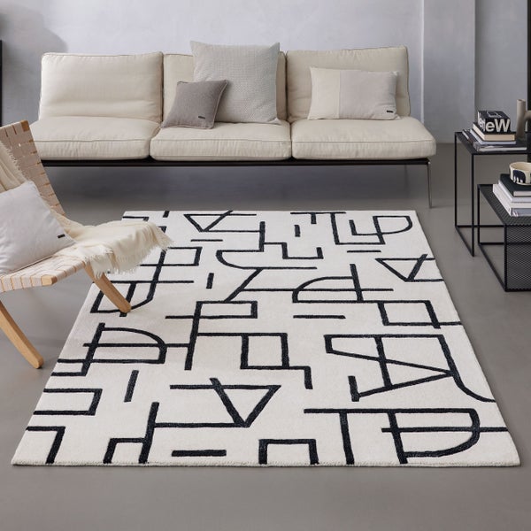 Moderner rechteckiger Teppich mit abstraktem schwarzen Muster in einem hellen Wohnzimmer mit Sofa und Sessel aus Holz.
