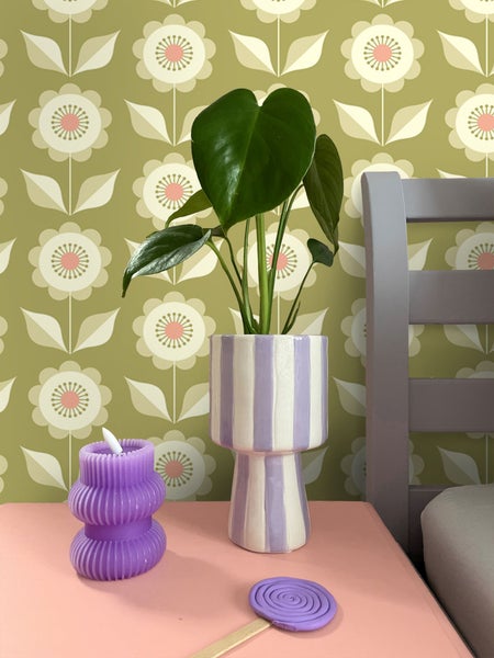 Dekorative Szene mit Zimmerpflanze im gestreiften Topf, Kerze und Bett auf einem Tisch vor einer gemusterten Tapete