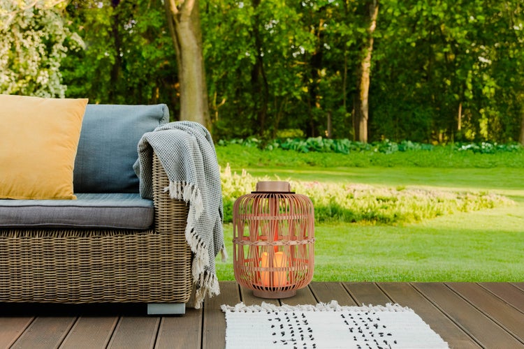 Szene mit Korblaterne, Kissen und Decke auf einer Terrasse mit Gartenblick