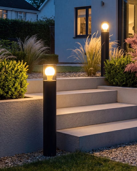 Zwei moderne, schwarze Pollerleuchten beleuchten Betontreppen und Pflanzen in einem Garten bei Dämmerung.