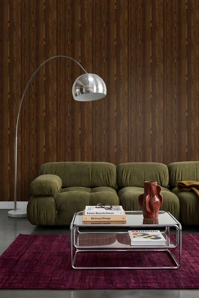 Wohnzimmer mit dunkler Holzoptik Tapete, beigem Sofa, Bogenlampe und einem Glastisch auf einem weinroten Teppich.