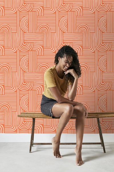 Frau sitzt vor einer Wand mit einem geometrischen Muster aus Linien und Bögen.
