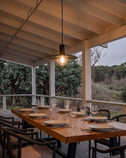 Gedeckter Esstisch aus Holz auf einer überdachten Terrasse mit geflochtenen Stühlen und hängender Außenleuchte im Abendlicht.