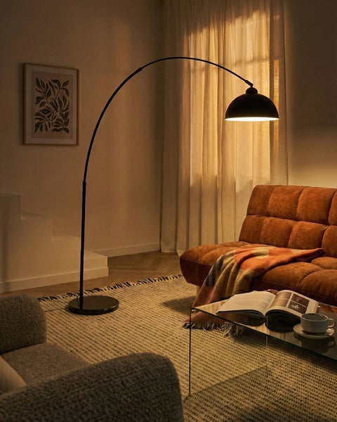 Moderne Wohnzimmerszene mit schwarzer Bogenstehleuchte, rostfarbenem Sofa, Glastisch und gewebtem Teppich in warmer Atmosphäre.