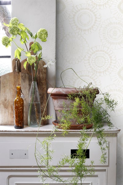 Dekorative Anordnung mit Blumen, Flaschen und Topfpflanze auf einem Schrank mit Blumendesign-Tapete.