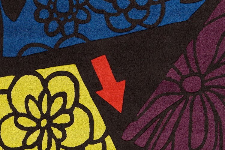 Detailansicht einer gemusterten Fußmatte mit Blumenmotiven in Gelb, Blau und Violett sowie einem roten Pfeil.