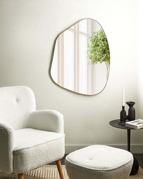Wandspiegel in asymmetrischer, organischer Form mit schmalem Metallrahmen in einem hellen Wohnraum neben einem weißen Sessel.