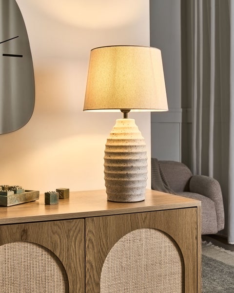 Tischleuchte mit strukturiertem Keramikfuß und Stoffschirm auf einem Sideboard aus Holz und Geflecht in einem hellen, aufgeräumten Raum.