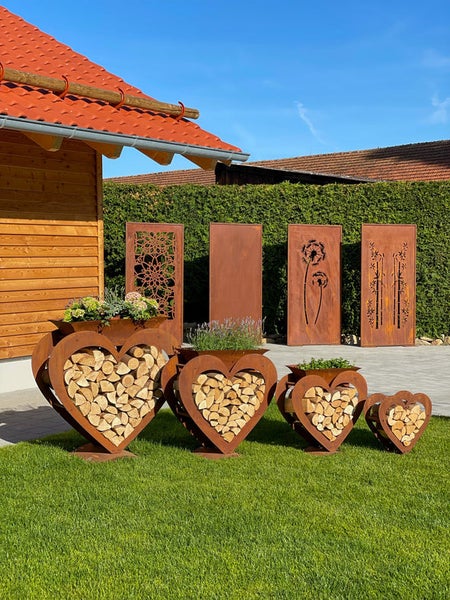 Vier herzförmige Kaminholzregale aus Edelrost mit integrierten Pflanzgefäßen und dekorative Sichtschutzwände aus Metall in einem Garten.