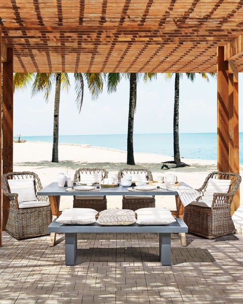 Gedeckter Esstisch mit Rattanstühlen und Holzboden unter einer hölzernen Pergola mit Blick auf den Strand