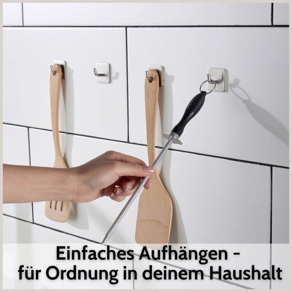 Selbstklebende Wandhaken an einer weißen Fliesenwand zur Aufbewahrung von Küchenutensilien wie Holzspateln und einem Wetzstab.