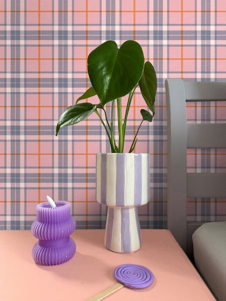 Monstera Pflanze im gestreiften Übertopf neben violetter Kerze vor karierter Tapete.