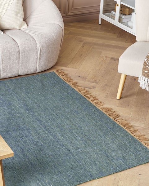 Blauer Flachgewebe-Teppich mit Fransen auf Fischgrätparkett in einem hell eingerichteten Wohnzimmer mit Sessel und weißem Schrank.