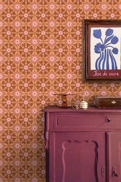 Tapete mit floralem Mandalamuster in Orange und Braun hinter einer violetten Kommode mit dekorativer Kunst und Accessoires.