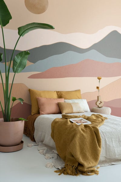 Schlafzimmer mit Wandtapete im Bergdesign in Erdtönen, Bett mit ockerfarbenen Kissen und großer Zimmerpflanze im Topf.