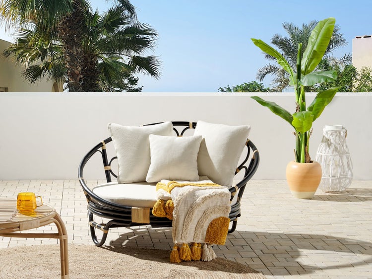 Terrassenszene mit Rattansessel, Kissen, Überwurf, Tisch und Pflanze für eine helle, organisierte Atmosphäre.