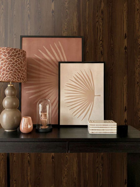 Wohnbereich mit Konsole, Tischlampe, zwei Wandbildern mit Palmenblattmotiven und Dekoration vor einer dunklen Holzoptik Tapete.