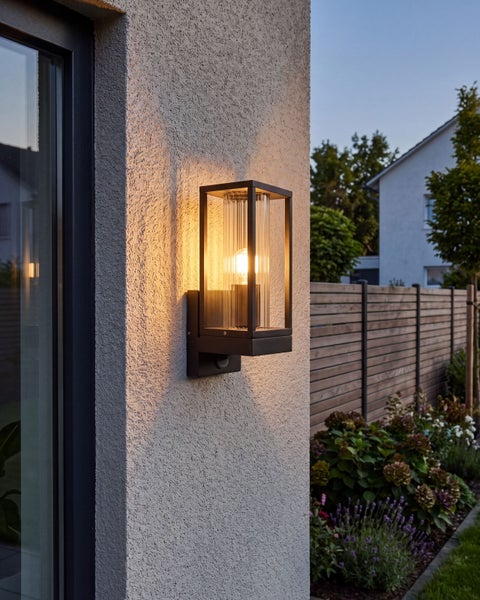 Moderne Außenwandleuchte mit Bewegungsmelder in rechteckiger Rahmenoptik montiert an einer Hausfassade im Garten