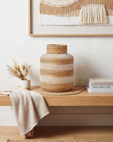 Dekorative Vase aus geflochtenem Naturmaterial auf einer Holzbank in einem hellen Wohnbereich mit Wohntextilien.