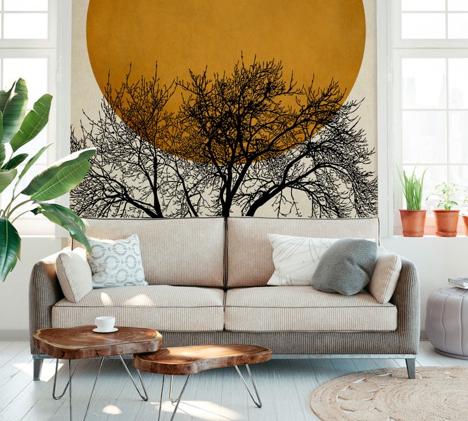 Fototapete mit Baumsilhouetten vor einer großen Sonne in einem hellen Wohnzimmer hinter einem beigen Sofa und Holztischen.