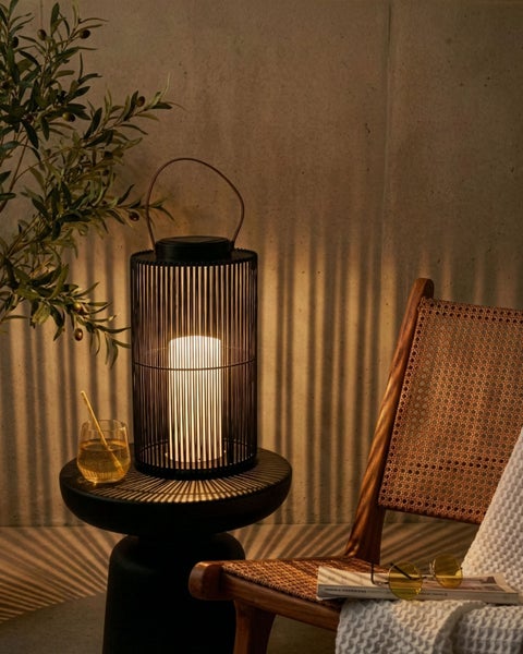 Moderne schwarze Solar-Laterne mit vertikalen Streben und integrierter LED-Lichtquelle auf einem Beistelltisch neben einem Rattan-Stuhl.