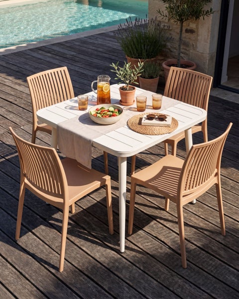 Modernes Garten-Set auf Holzterrasse mit weißem Tisch und vier beigen Stühlen mit Lamellenrücken neben einem Pool.