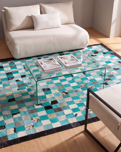 Moderner Flicken-Teppich mit türkisfarbenen, grauen und weißen Quadraten unter einem Couchtisch aus Glas, neben einem beigen Sofa.