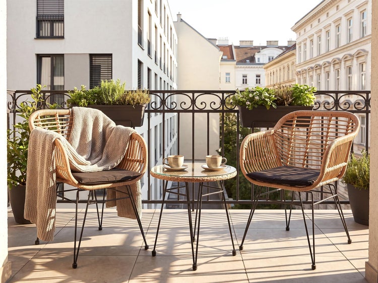 Gemütlicher Balkon mit zwei Rattan Sesseln, einem kleinen Beistelltisch, Sitzkissen, einer Decke und Balkonkästen vor einer städtischen Kulisse.