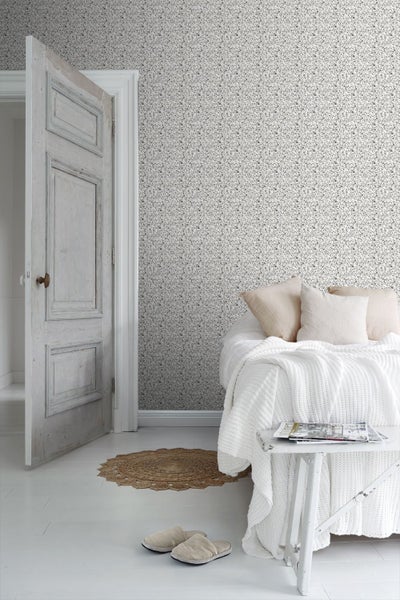 Helles Schlafzimmer mit kleinblumig gemusterter Tapete, einer offenen alten Holztür und einem Bett mit weißen Strickdecken.