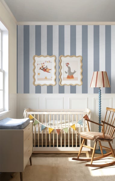 Helles Babyzimmer mit blau-weiß gestreifter Tapete, weißer Wandtäfelung, Holzbett mit Wimpelkette, einer Kommode und einem Holzschaukelstuhl.