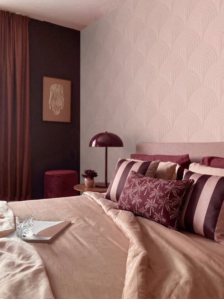 Schlafzimmer mit einer Tapete mit geometrischem Bogenmuster, einem Bett mit Dekokissen und einer Nachttischlampe.