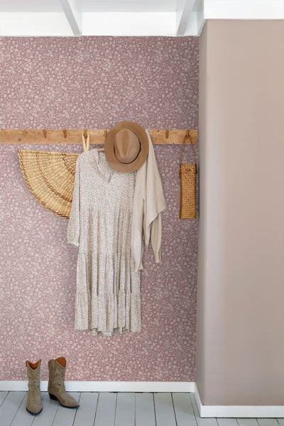 Vliestapete mit floralem Muster in Altrosa, kombiniert mit einer Garderobenleiste aus Holz, Hut und Kleid in einem hellen Raum.