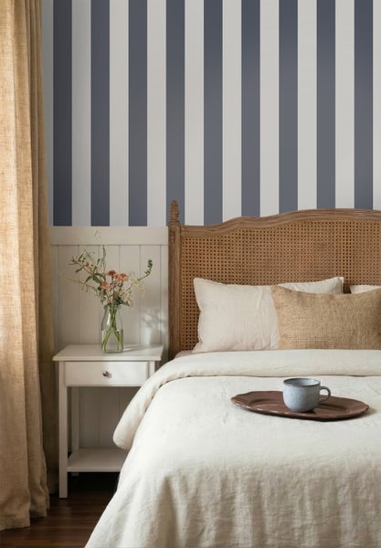 Helles Schlafzimmer mit vertikal gestreifter Tapete über weißer Wandtäfelung, Holzbett mit Rattankopfteil und Nachttisch.