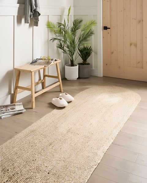 Ein heller Flur mit einem ovalen Läufer aus Naturfaser, einer Holzbank und Zimmerpflanzen in Töpfen vor einer Holztür.