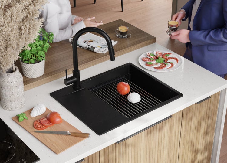 Schwarzes Spülbecken mit Armatur auf einer Küchentheke, die mit Tomaten und Mozzarella belegt ist. Zwei Personen unterhalten sich im Hintergrund.