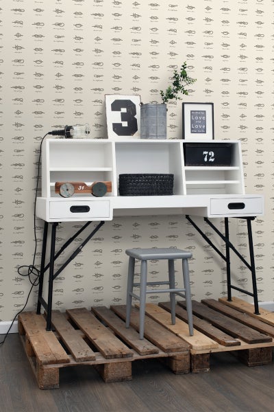 Szene mit weißem Schreibtisch, grauem Hocker und Dekorationsartikeln auf einer Holzpalette vor einer Wand mit Knotenmuster