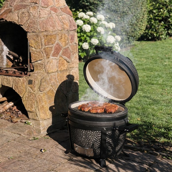 Schwarzer Keramik-Kamadogrill mit offenem Deckel und rauchenden Steaks auf dem Grillrost neben einem gemauerten Gartenkamin auf einer Terrasse.