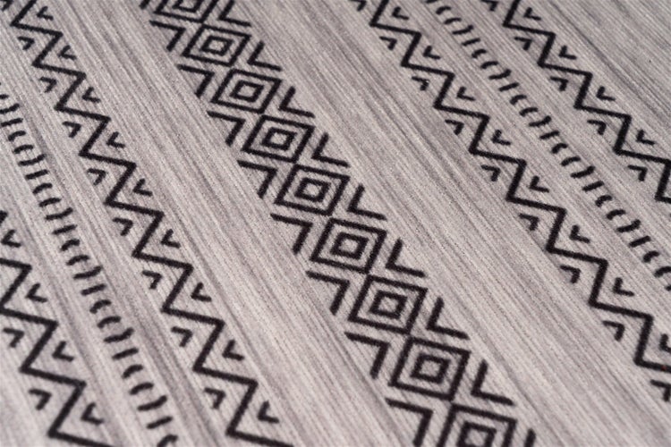 Nahaufnahme eines Teppichs mit schwarzem geometrischem Muster aus Zickzacklinien und Rauten auf grauem strukturiertem Stoff.