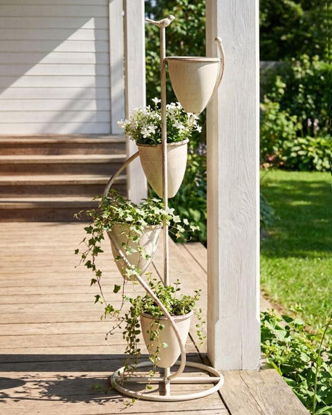 Blumenständer aus Metall mit vier Töpfen in Spiralform und Vogelfigur auf einer hellen Holzterrasse mit Blick in den Garten.
