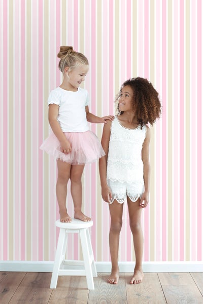 Zwei Mädchen in einem hellen Raum mit gestreifter Tapete, eines auf einem weißen Hocker.