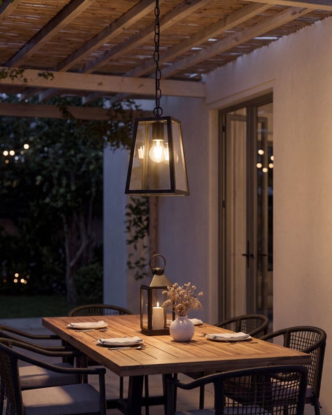 Moderne Hängeleuchte in Laternenform über einem hölzernen Esstisch auf einer abendlichen Terrasse mit Stühlen und Tischdekoration.
