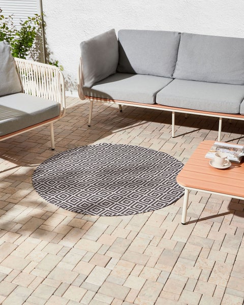 Runder Outdoor-Teppich mit geometrischem Rautenmuster in Schwarz-Weiß auf einer hellen Steinterrasse mit modernen Gartenmöbeln.