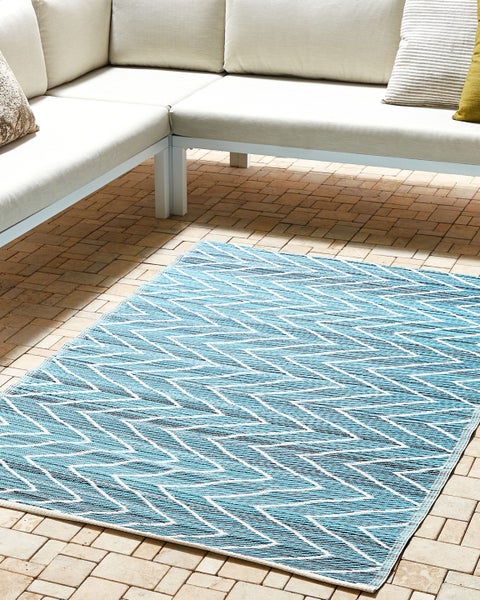 Outdoor-Teppich mit blau-weißem Zickzack-Muster auf einer hellen Terrasse mit Fliesenboden vor einem modernen Ecksofa.