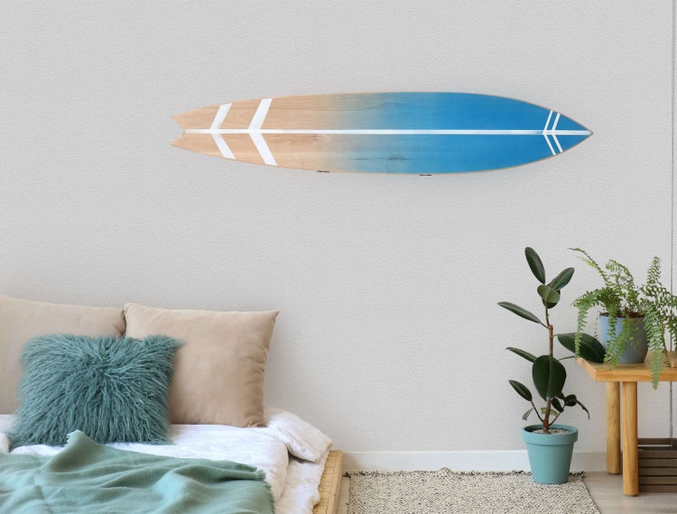 Dekorative Wanddekoration Surfbrett aus Holz mit blauem Farbverlauf und weißen Streifen über einem Bett mit Kissen und Zimmerpflanzen.